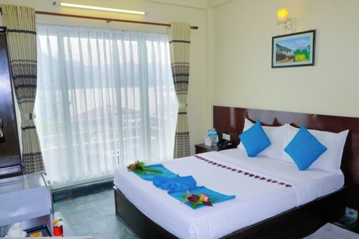 Una habitación de hotel con una cama, cortinas, una mesita de noche, una lámpara y un cuadro en la pared.