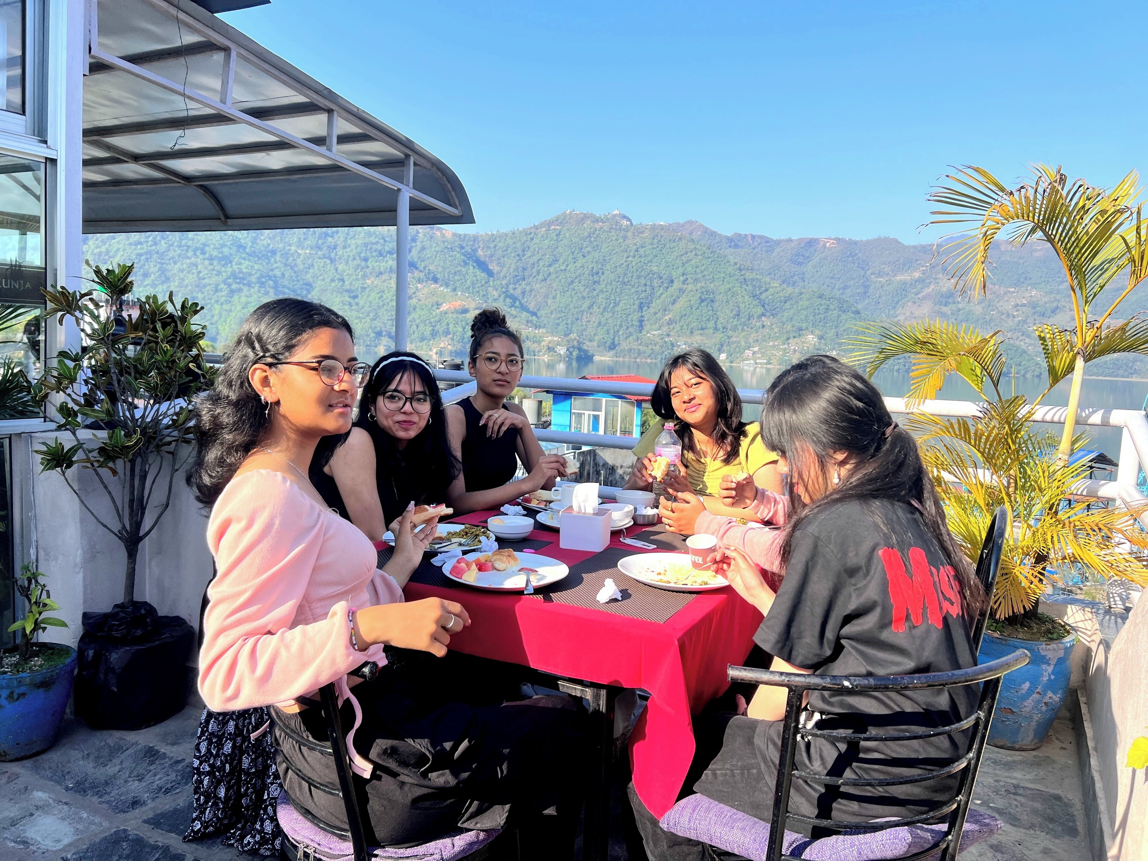 Un grupo de personas cenando en una mesa al aire libre con mantel rojo, rodeado de plantas en macetas y con vistas a un paisaje montañoso.