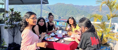 Un grupo de personas cenando en una mesa al aire libre con mantel rojo, rodeado de plantas en macetas y con vistas a un paisaje montañoso.