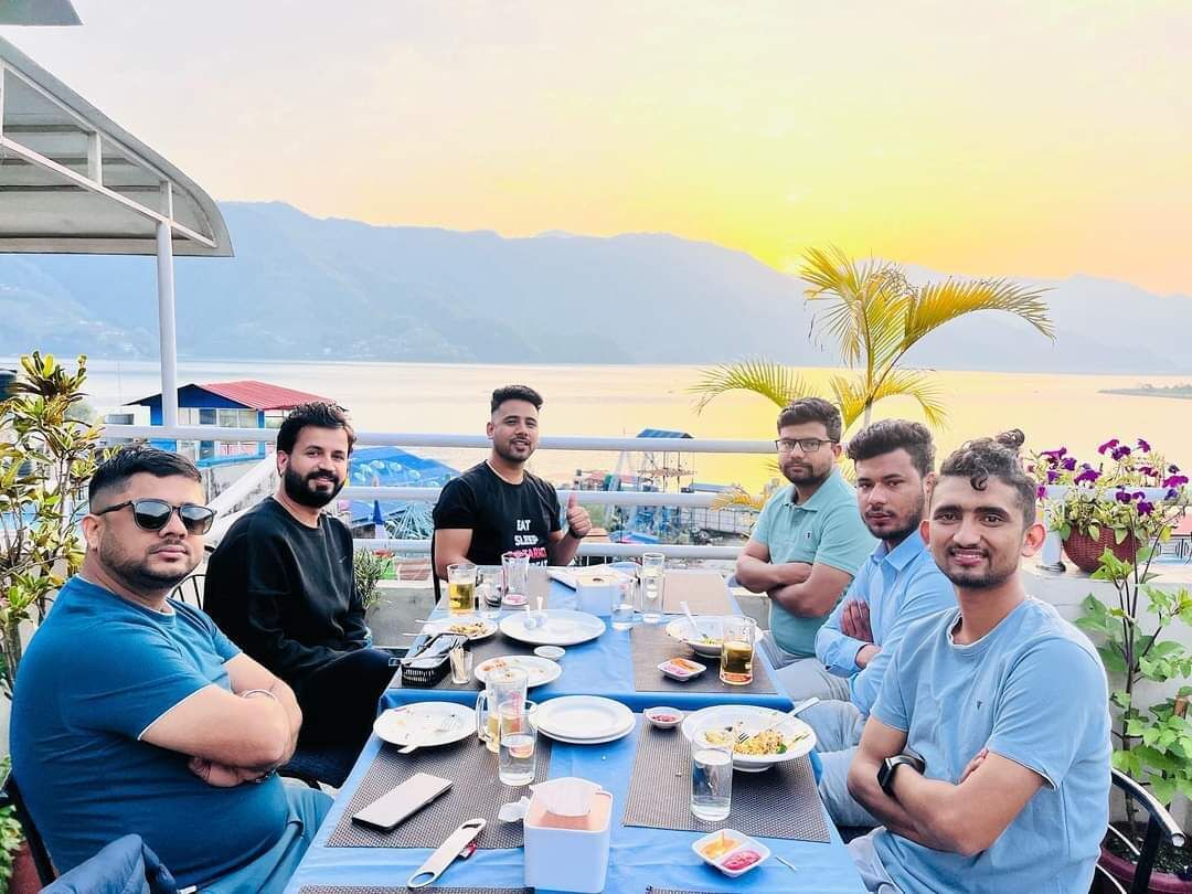 A group of people dining at an outdoor table with a view of a body of water and mountains in the background.
