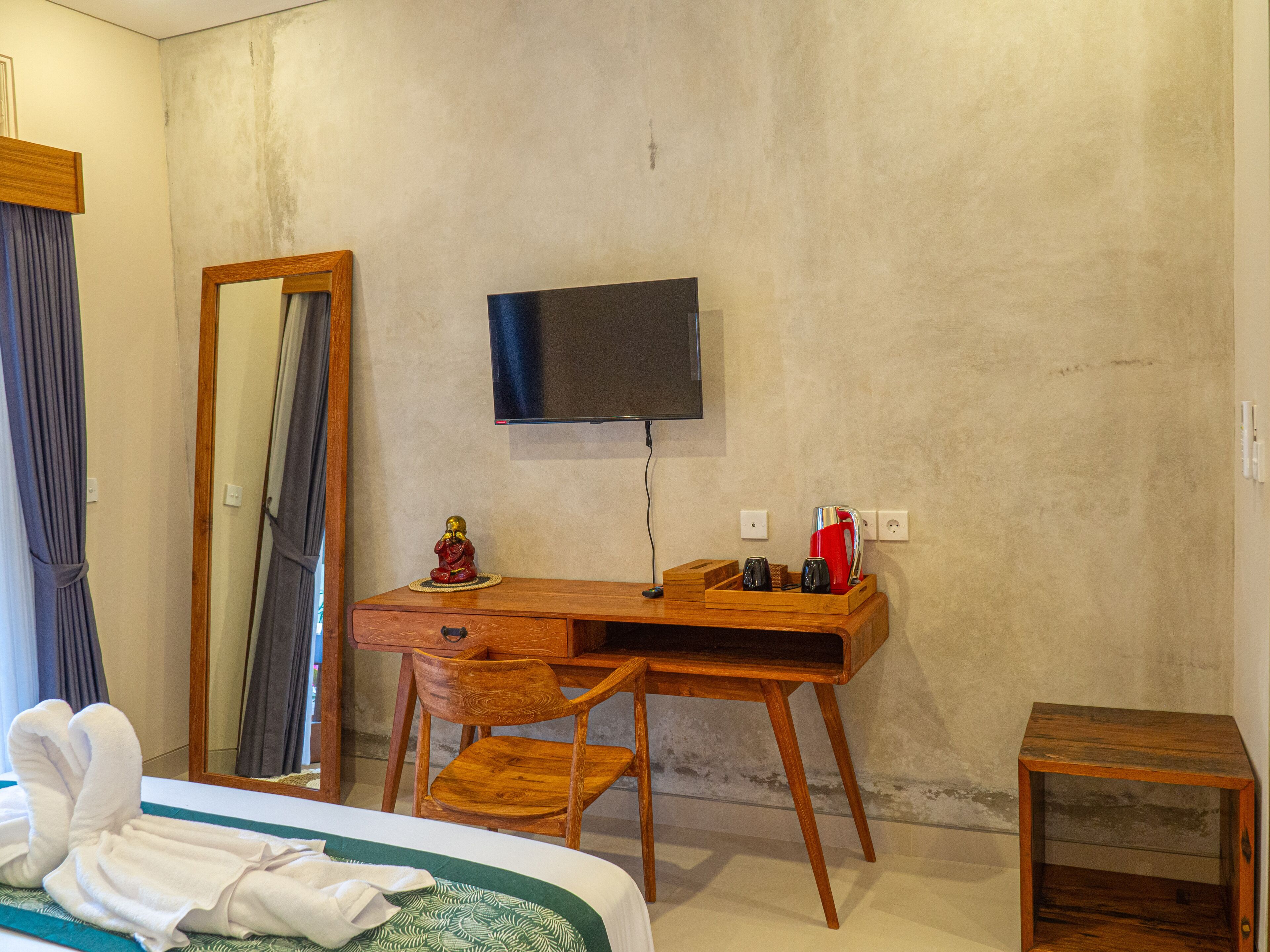 A hotel room with a wooden desk, a chair, a bed with a green and white patterned bedspread, a wooden nightstand, a television mounted on the wall, and a mirror.
