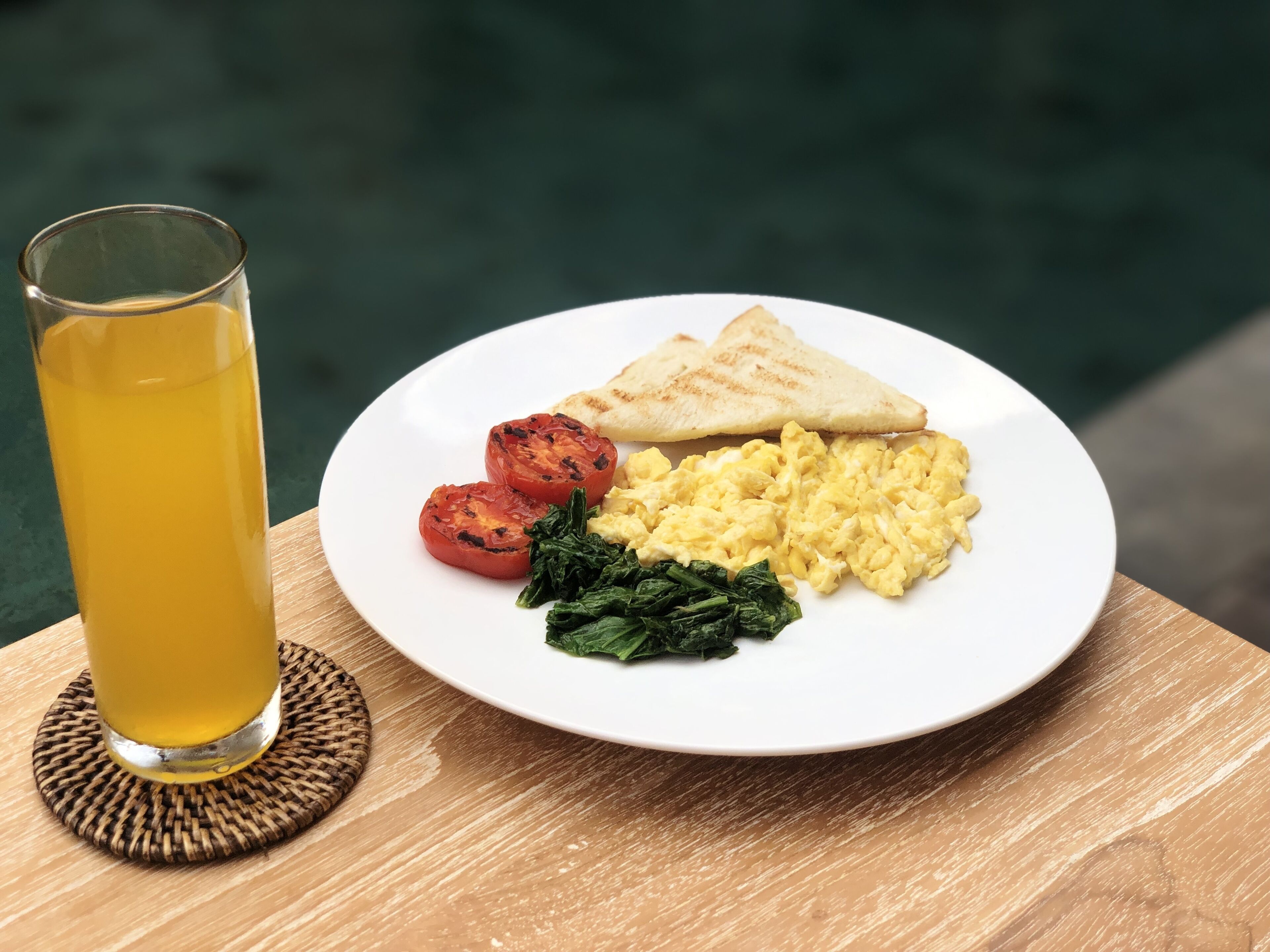 Ein Teller mit Rührei, gegrillten Tomaten, Spinat und Toast auf einem Holztisch, dazu ein Glas Orangensaft.