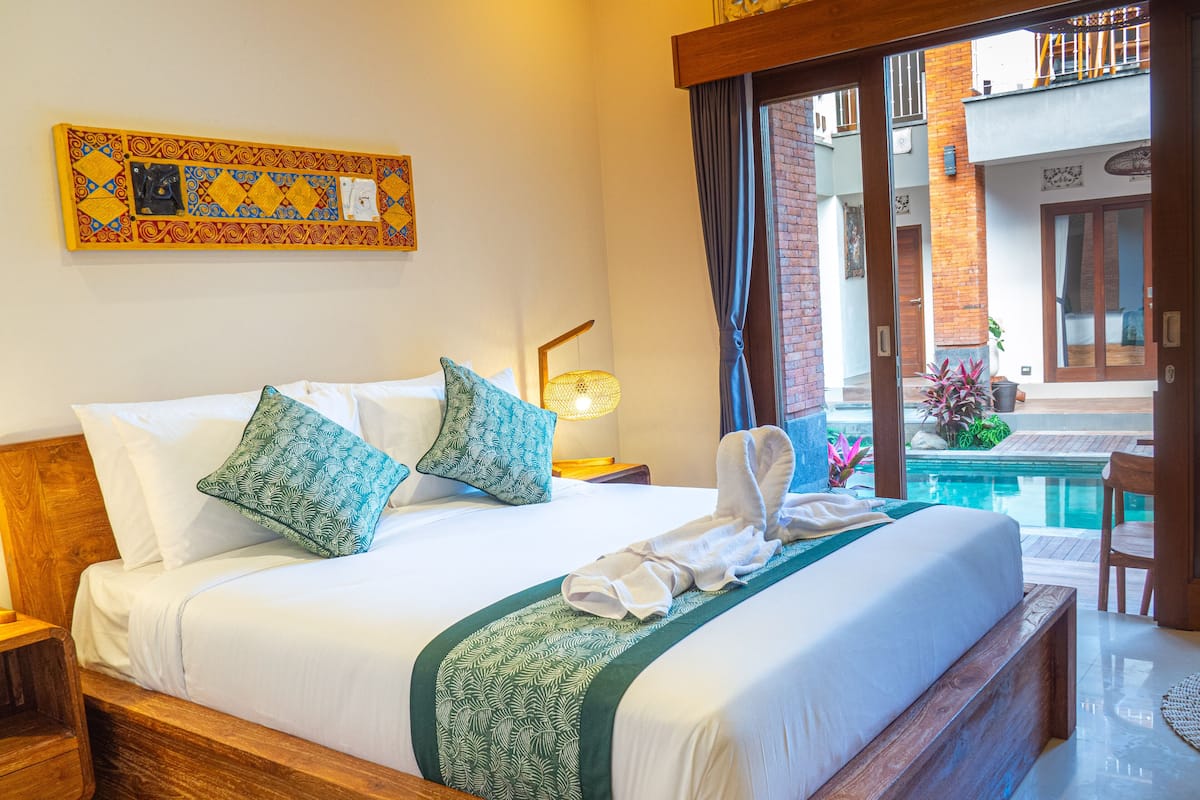 A neatly made bed with white linens and green patterned pillows, a wooden headboard, a small bedside table with a lamp, and a view of a swimming pool through a glass door.
