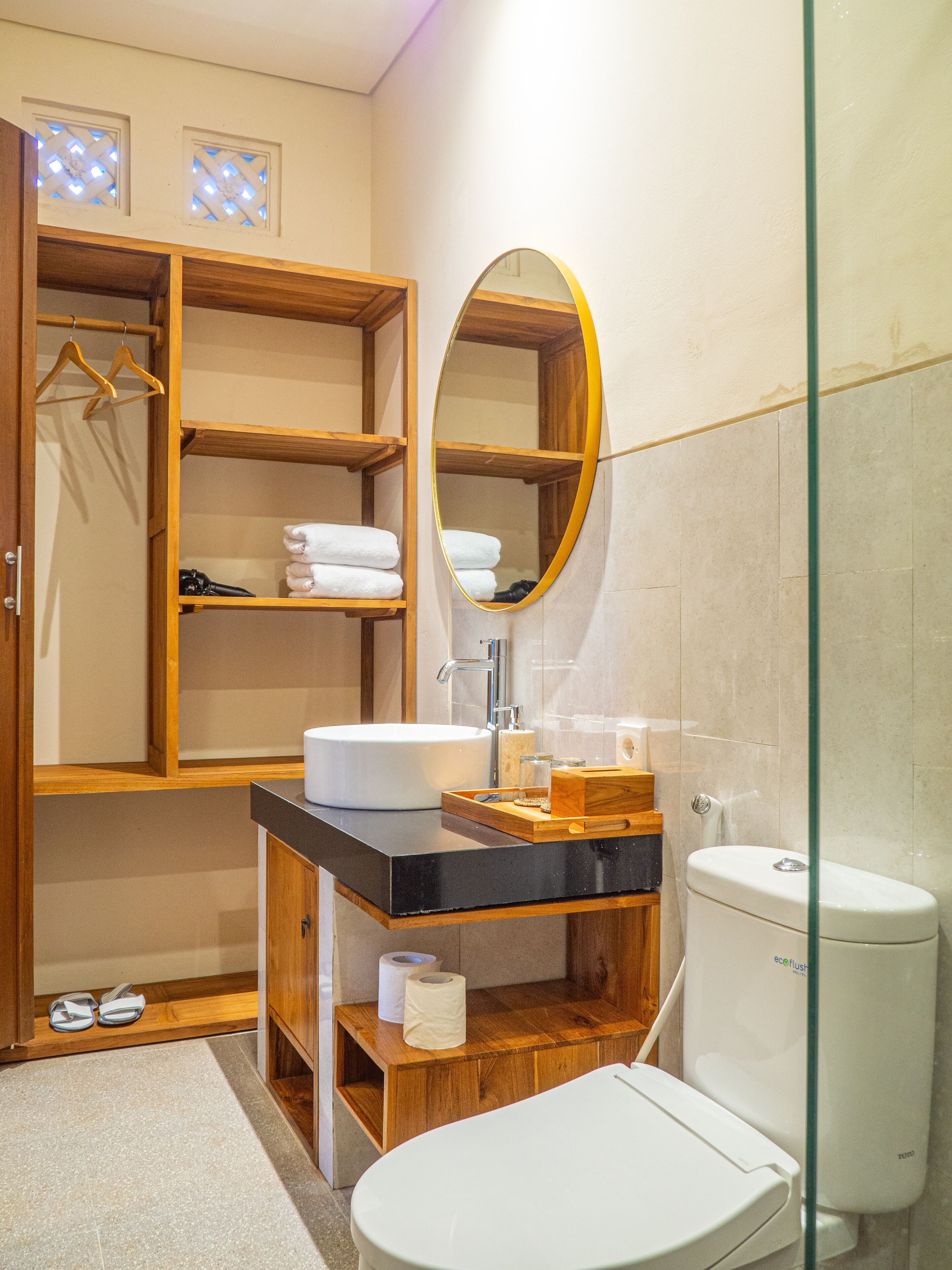 Une salle de bain moderne avec un meuble-lavabo en bois, un miroir rond, des toilettes blanches et une douche à parois de verre.