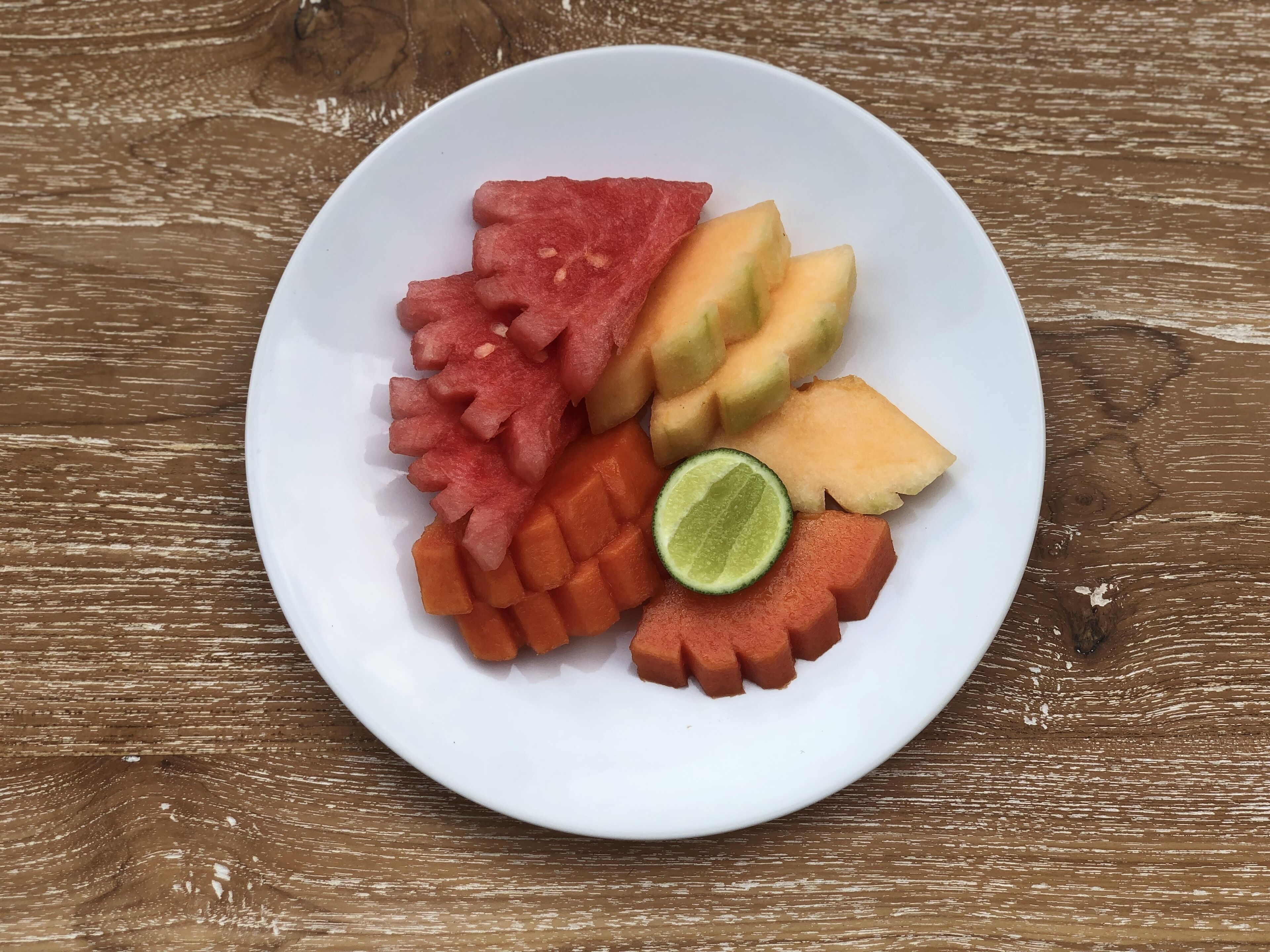 A white plate with various fruits including watermelon, cantaloupe, and lime on a wooden surface.