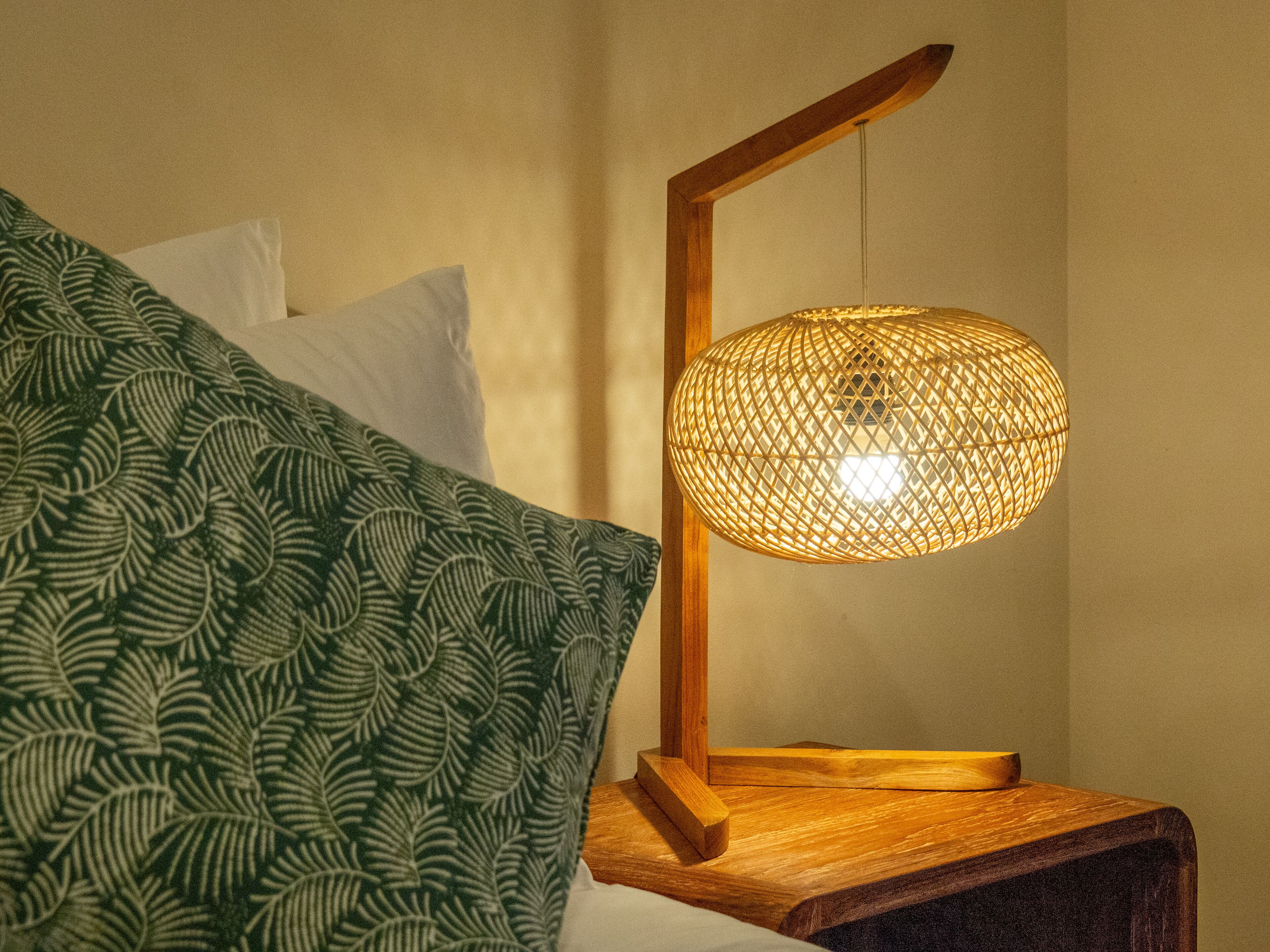 A bed with a green patterned blanket and a wooden bedside table with a woven lamp.