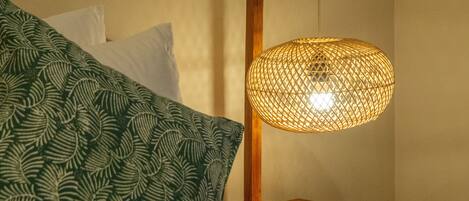 A bed with a green patterned blanket and a wooden bedside table with a woven lamp.