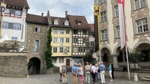 Eine Gruppe von Personen steht auf einem Kopfsteinpflasterplatz, umgeben von historischen Gebäuden, darunter ein Gebäude mit einer Schweizer Flagge.
