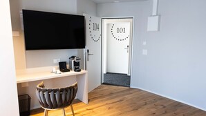 Ein modernes, minimalistisches Zimmer mit Holzboden, einem weißen Schreibtisch, einem Flachbildfernseher, einem gepolsterten Stuhl und zwei Türen mit den Nummern 101 und 104.