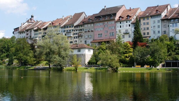 Eine Reihe mehrstöckiger Gebäude unterschiedlicher Architektur, ein stiller See im Vordergrund und eine kleine Parkanlage mit Bänken und Bäumen.