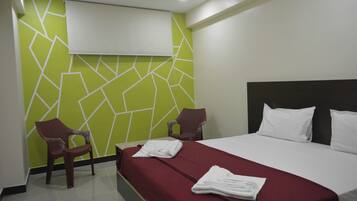 Habitación de hotel con cama, dos sillas, colcha roja, almohadas blancas y diseño geométrico verde en la pared.