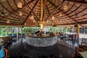 A spacious bar area with a central island, surrounded by seating and a high ceiling with woven thatch.