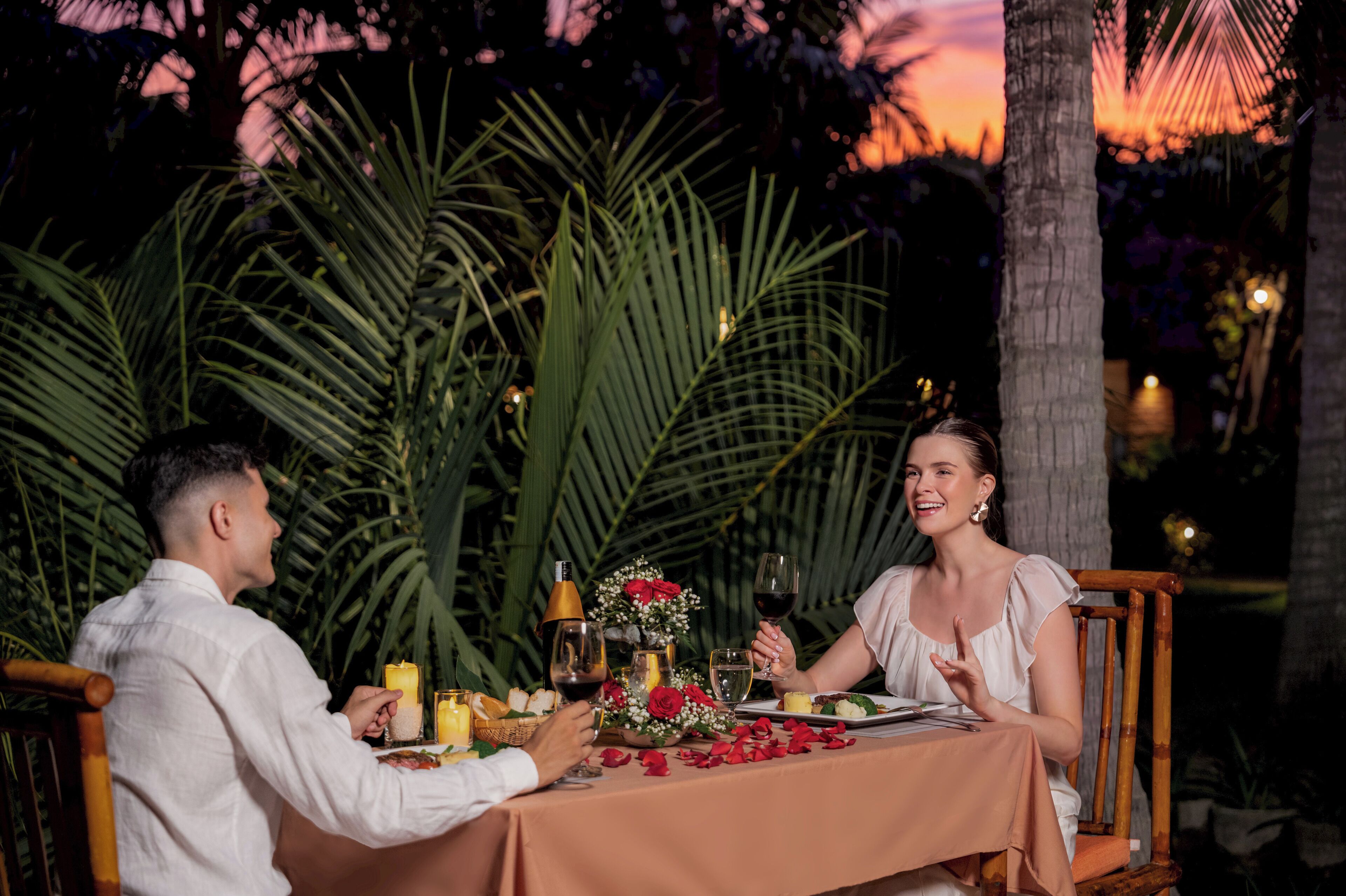 A couple dining outdoors at night with a table set for two, surrounded by greenery and a sunset in the background.
