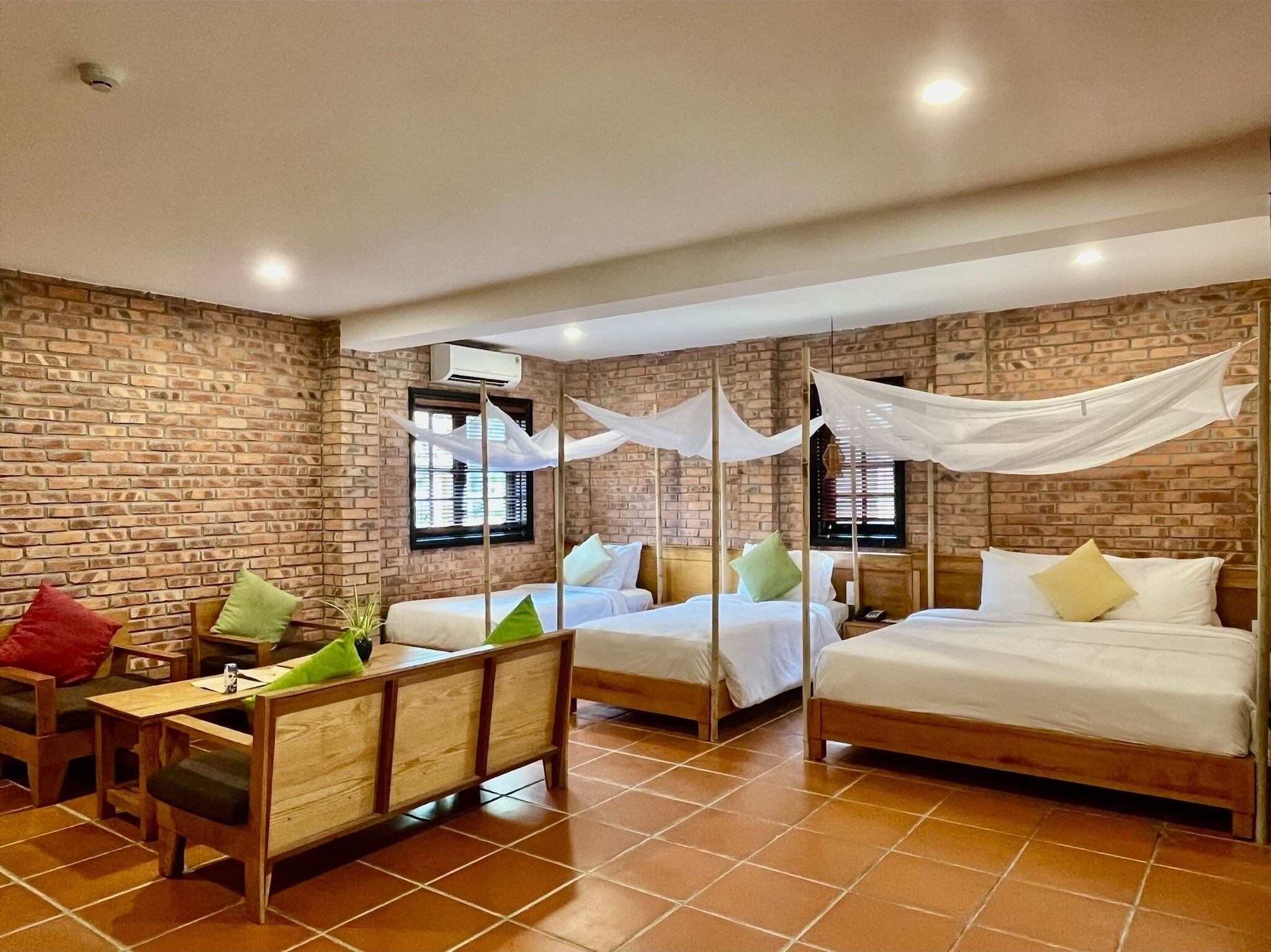 A hotel room with brick walls, two beds with white bedding, a wooden bench with cushions, and a small table.