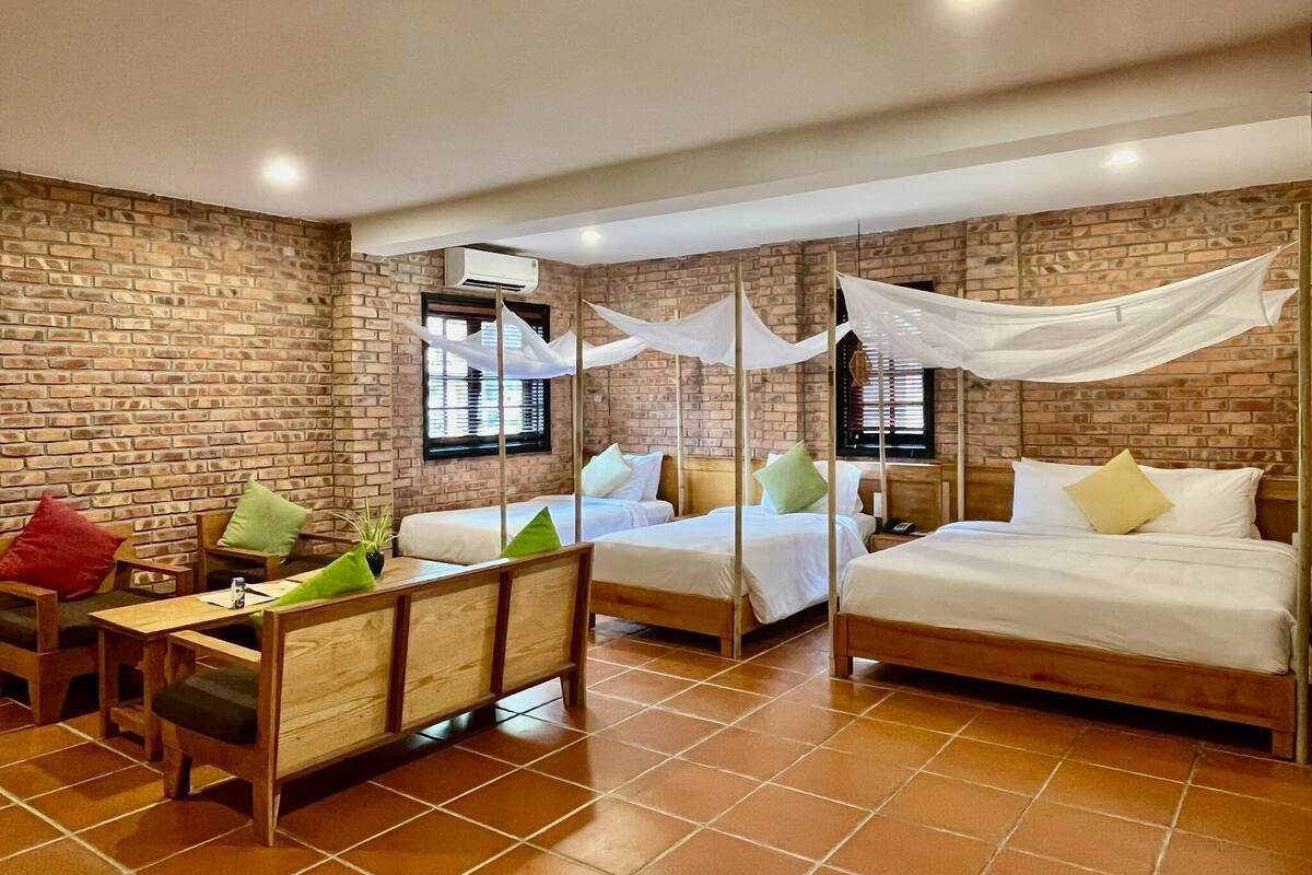 Une chambre d’hôtel avec des murs en briques, deux lits avec du linge de lit blanc, un banc en bois avec des coussins et une petite table.
