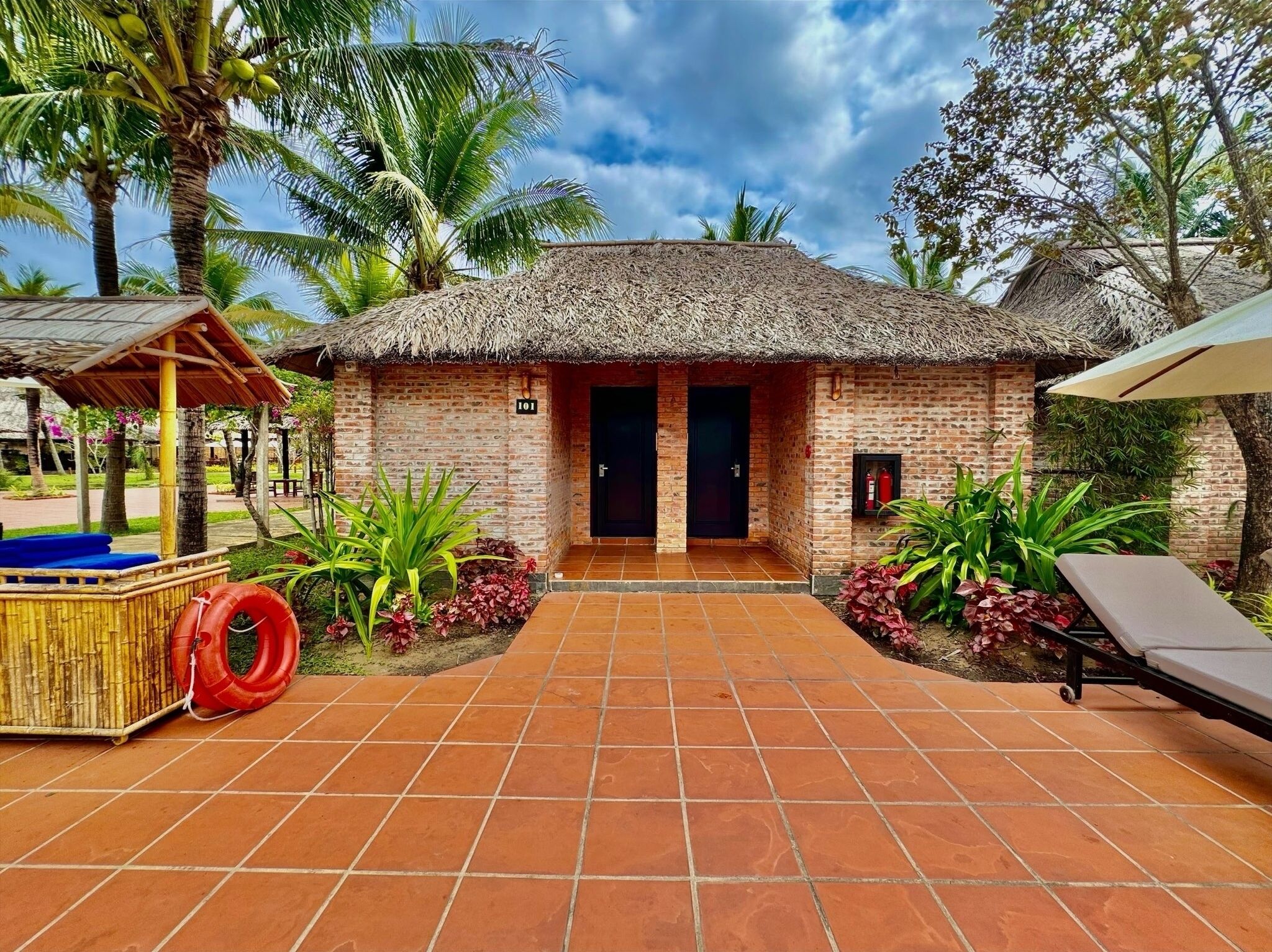 A thatched-roof building with a brick facade, a tiled patio, and a lounge chair.