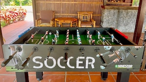 Une table de baby-foot portant l’inscription « SOCCER » et comportant plusieurs joueurs, placée dans une pièce avec un mur en bambou et une petite table en bois.