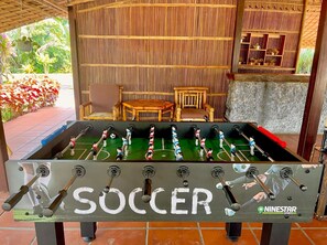 Ein Kicker mit der Aufschrift „SOCCER“ und mehreren Spielern, platziert in einem Raum mit Bambuswand und einem kleinen Holztisch.
