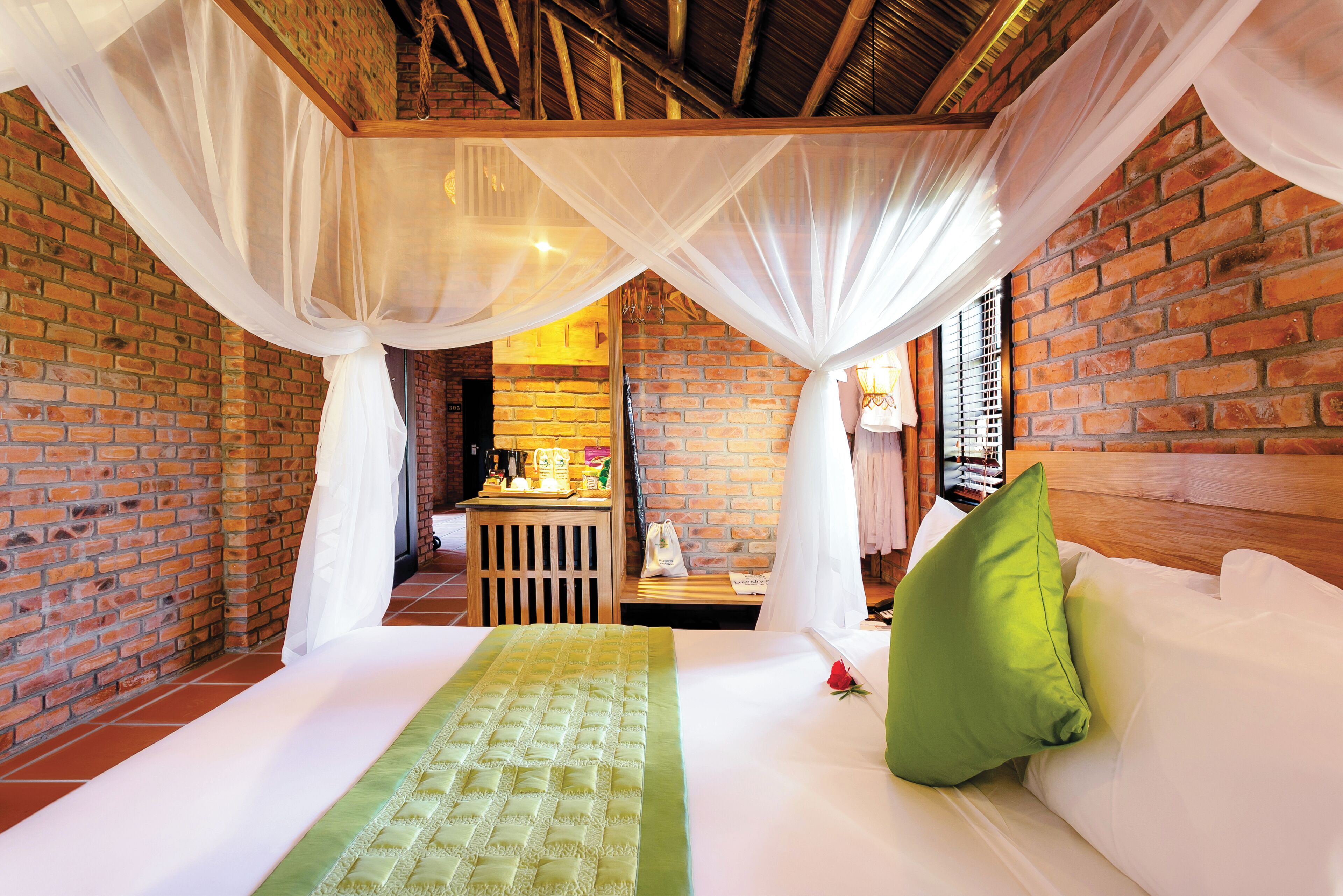 A room with a bed, a green bedspread, white curtains, and a brick wall.