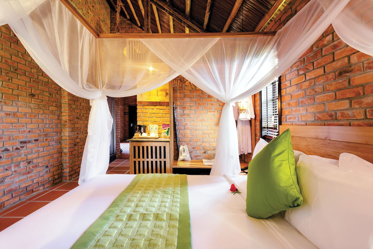 A room with a bed, a green bedspread, white curtains, and a brick wall.