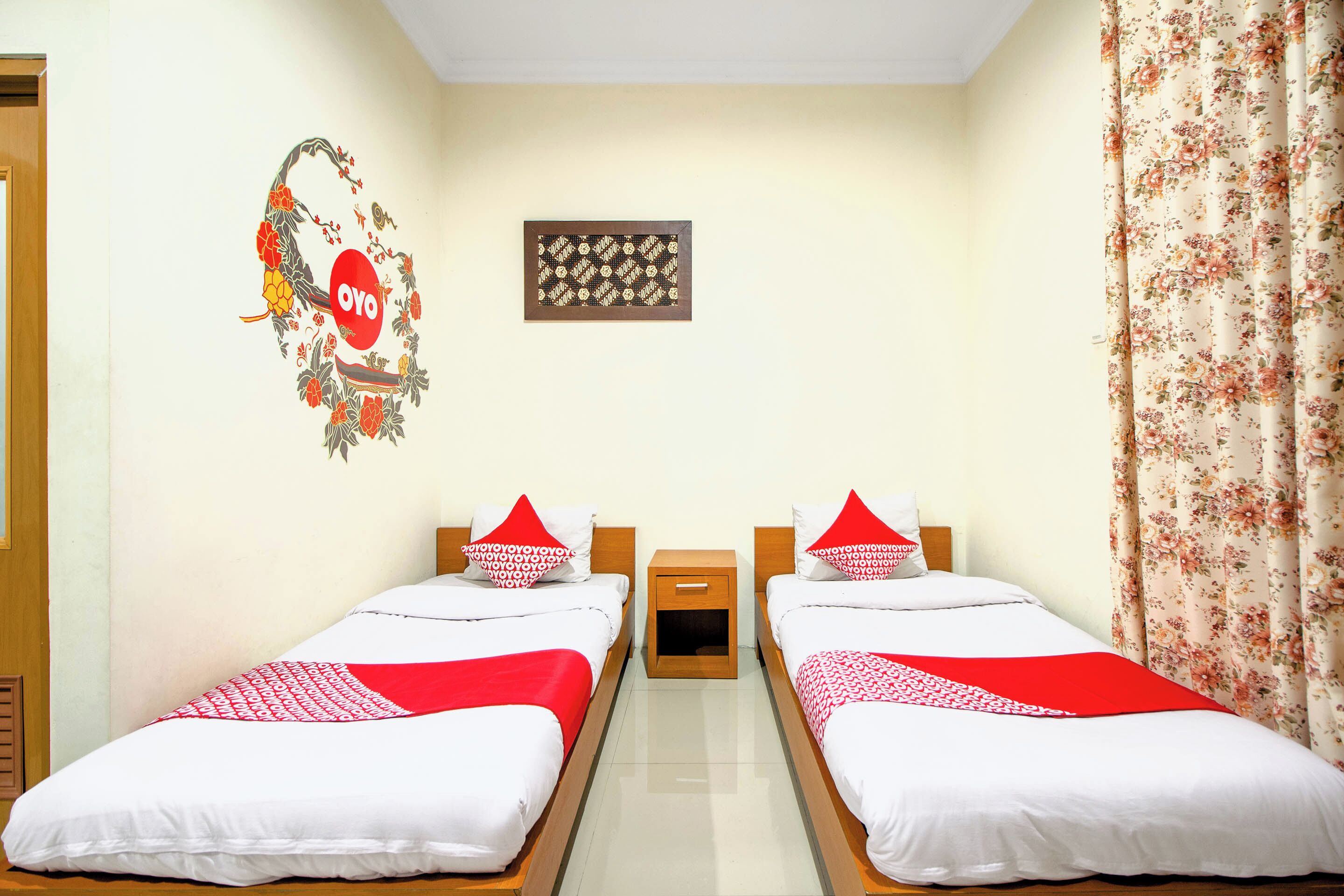 A hotel room with two beds, a wooden nightstand, a wall decoration, and a floral curtain.
