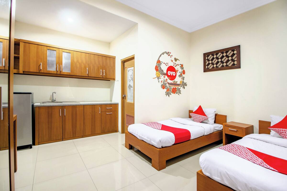 Habitación de hotel compacta con dos camas individuales, un pequeño armario de madera, un refrigerador y una cocina con lavamanos y alacenas.