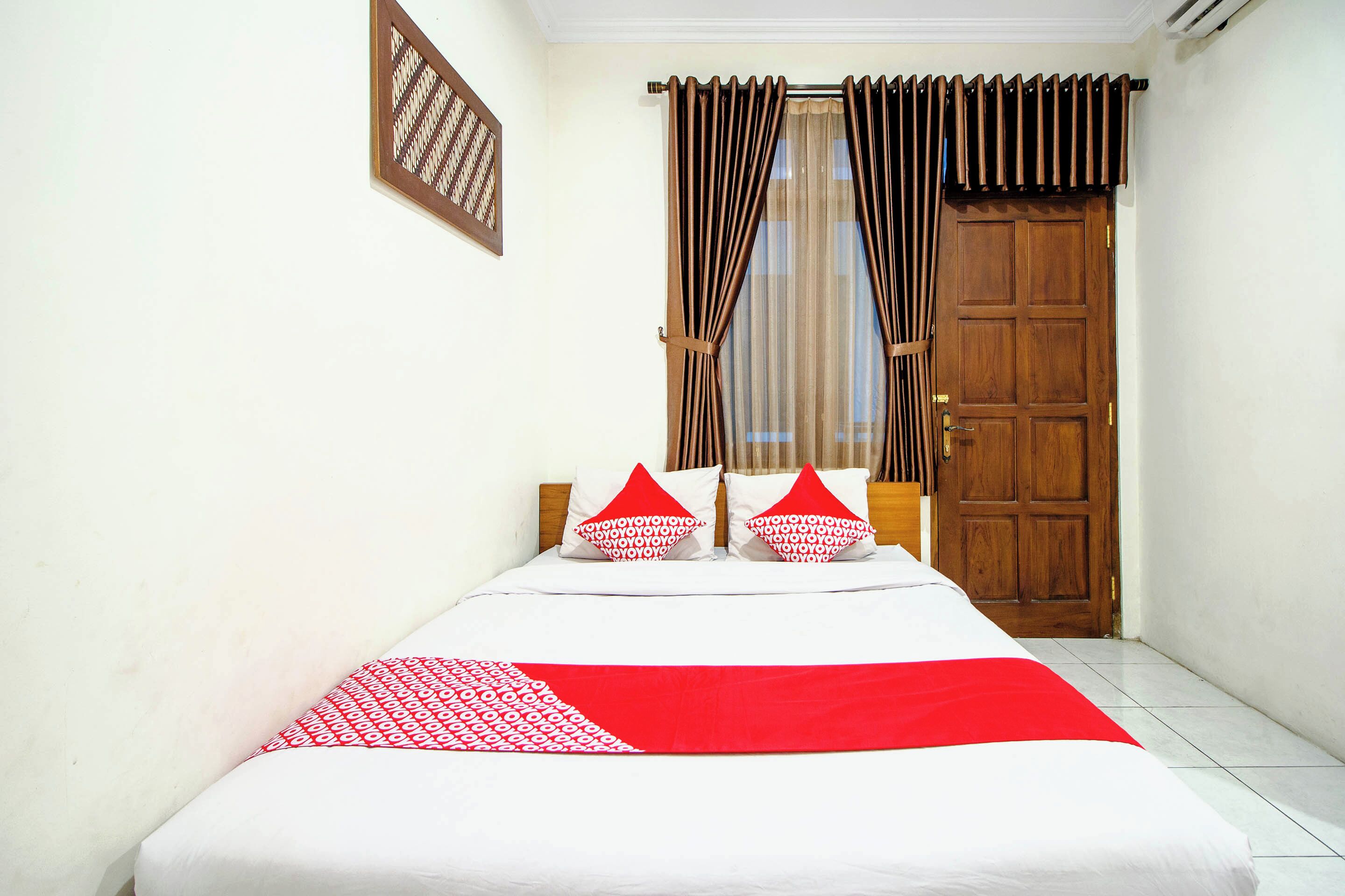 Une chambre à coucher avec un lit, deux oreillers, une couvre-lit rouge et blanche, une porte en bois et une fenêtre avec des rideaux.