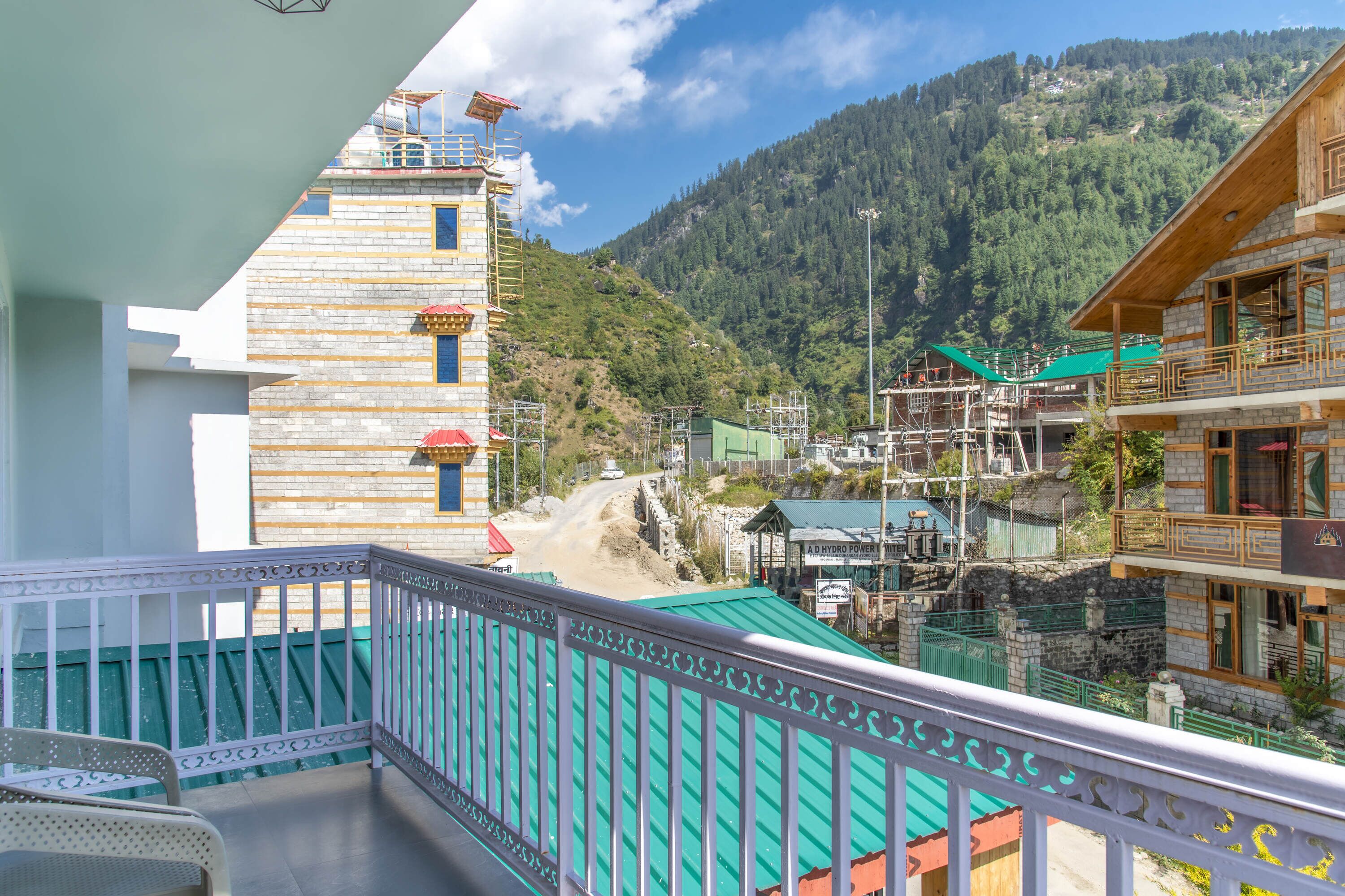 Un balcon donnant sur un paysage montagneux, un bâtiment portant une enseigne indiquant « A.D. HYDRO POWER », et un chantier de construction en arrière-plan.