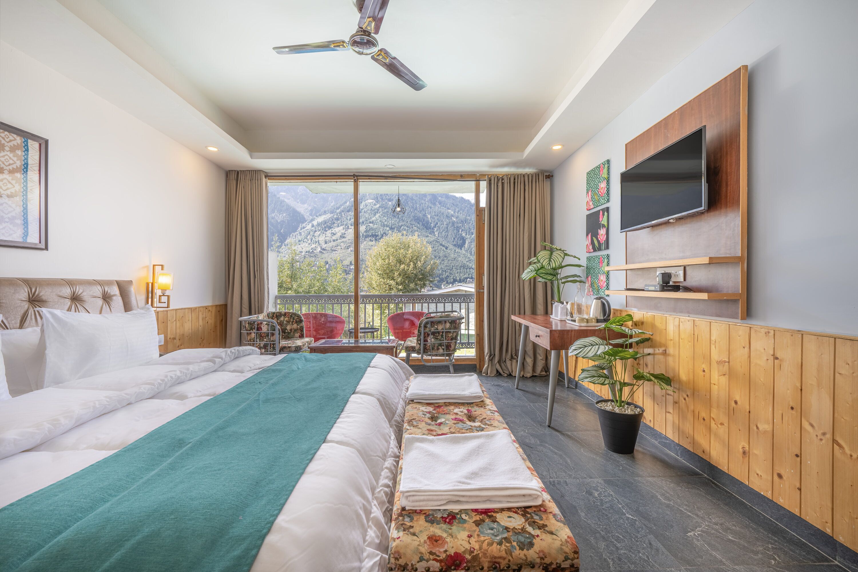 A hotel room with a large bed, a desk with a TV, a balcony with a view of mountains, and a ceiling fan.