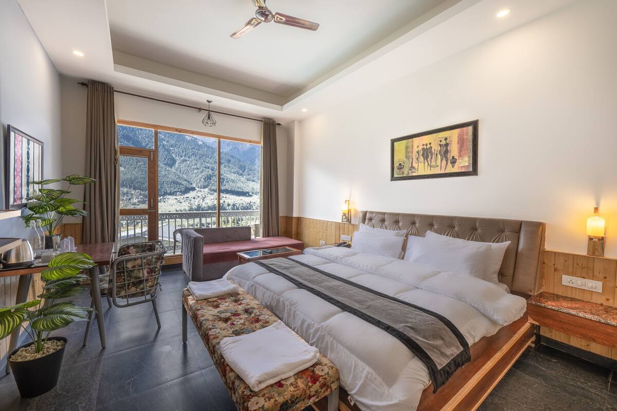 Habitación de hotel moderna con una cama grande, un escritorio con silla, un banco y vistas a montañas nevadas.