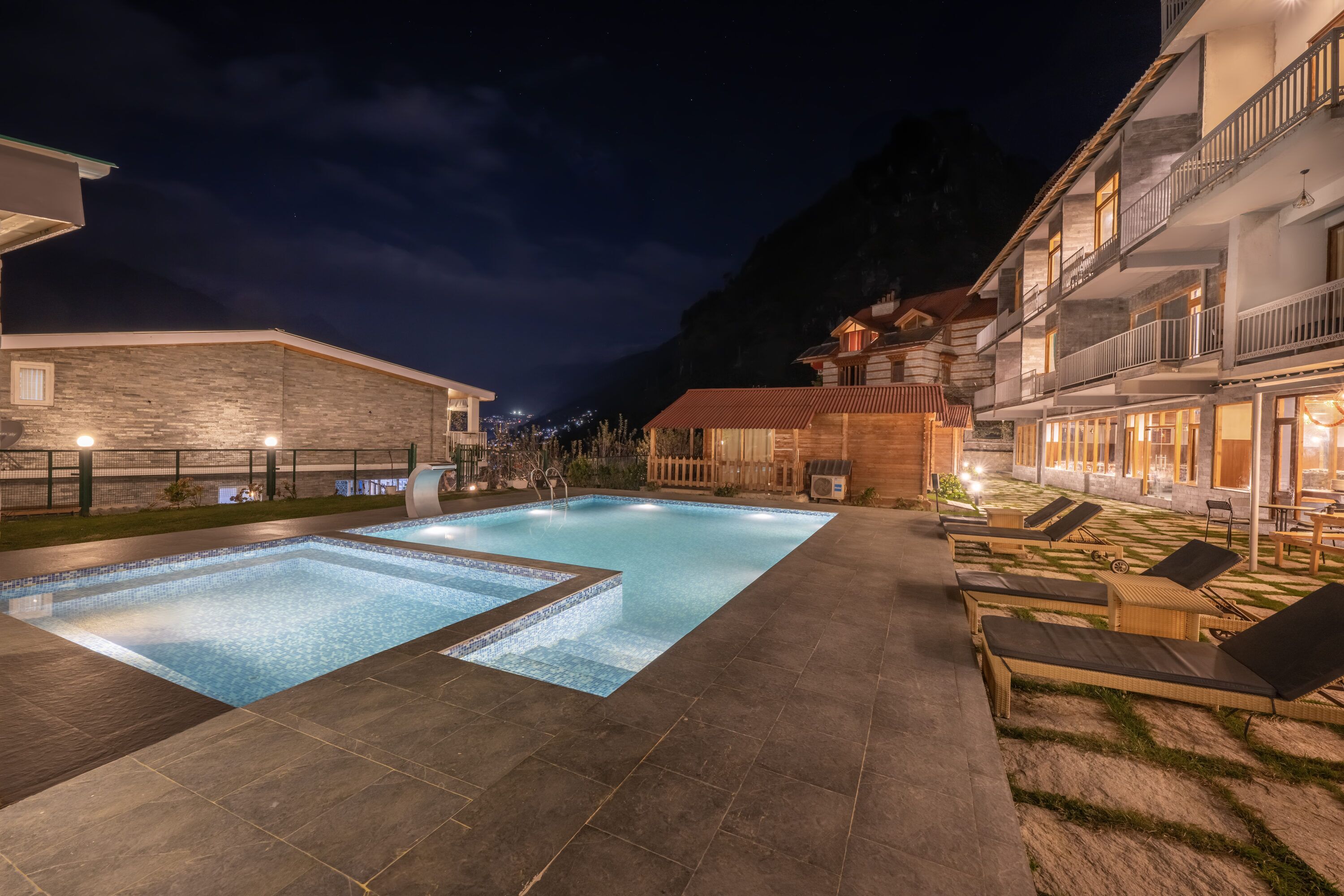A swimming pool with lounge chairs, a stone building, and a wooden structure with balconies.
