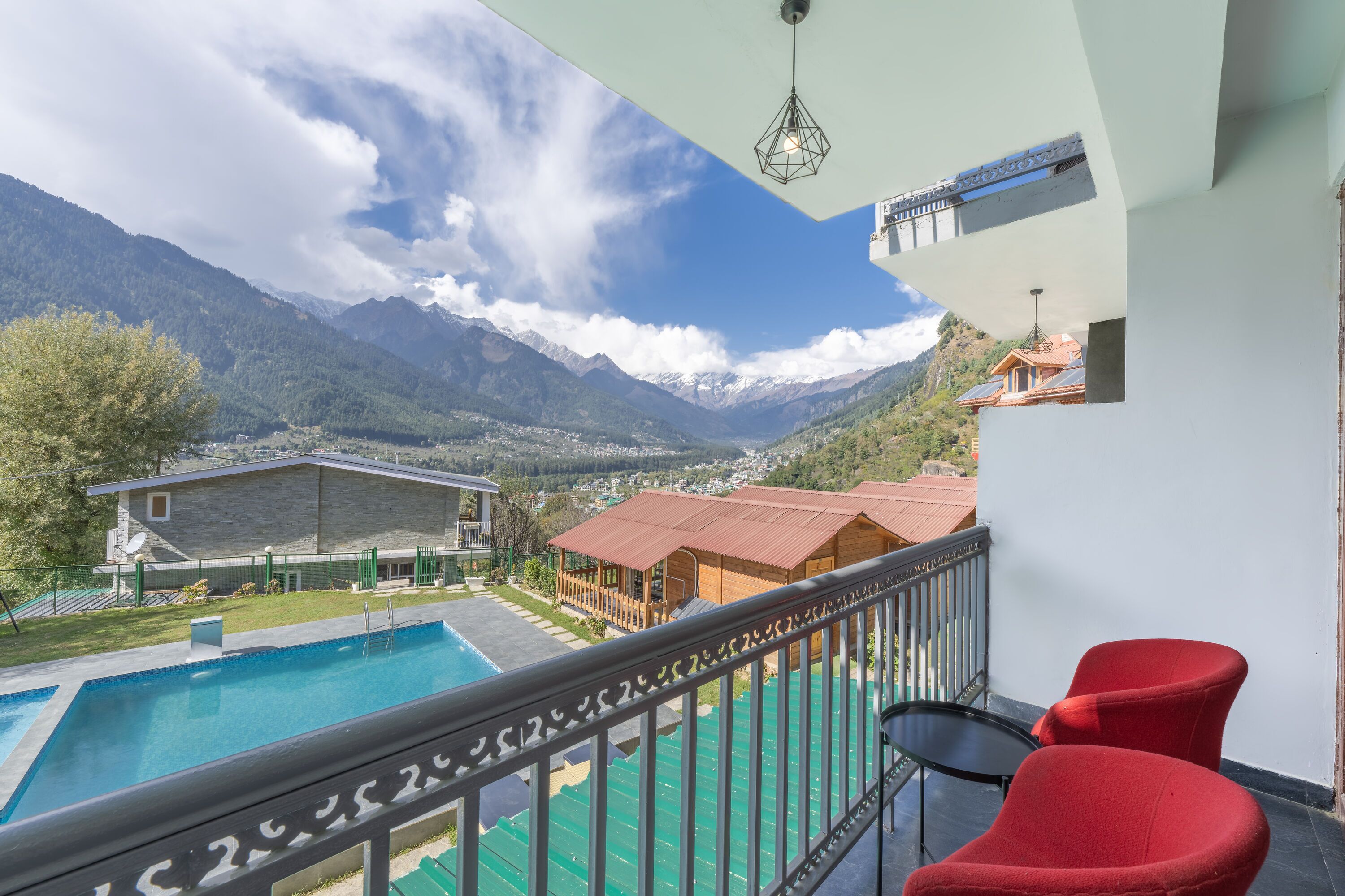 A balcony with a pool, red chairs, and a view of mountains and houses.