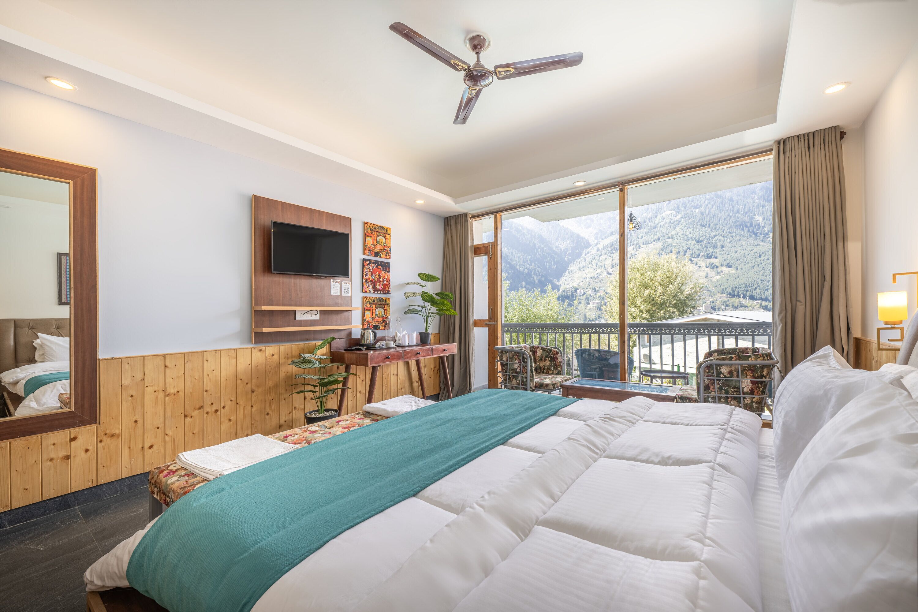 A hotel room with a large bed, a desk with a flat-screen TV, a ceiling fan, and a view of mountains through the window.