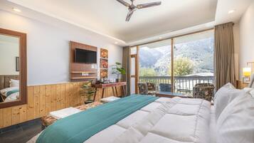 A hotel room with a large bed, a desk with a flat-screen TV, a ceiling fan, and a view of mountains through the window.