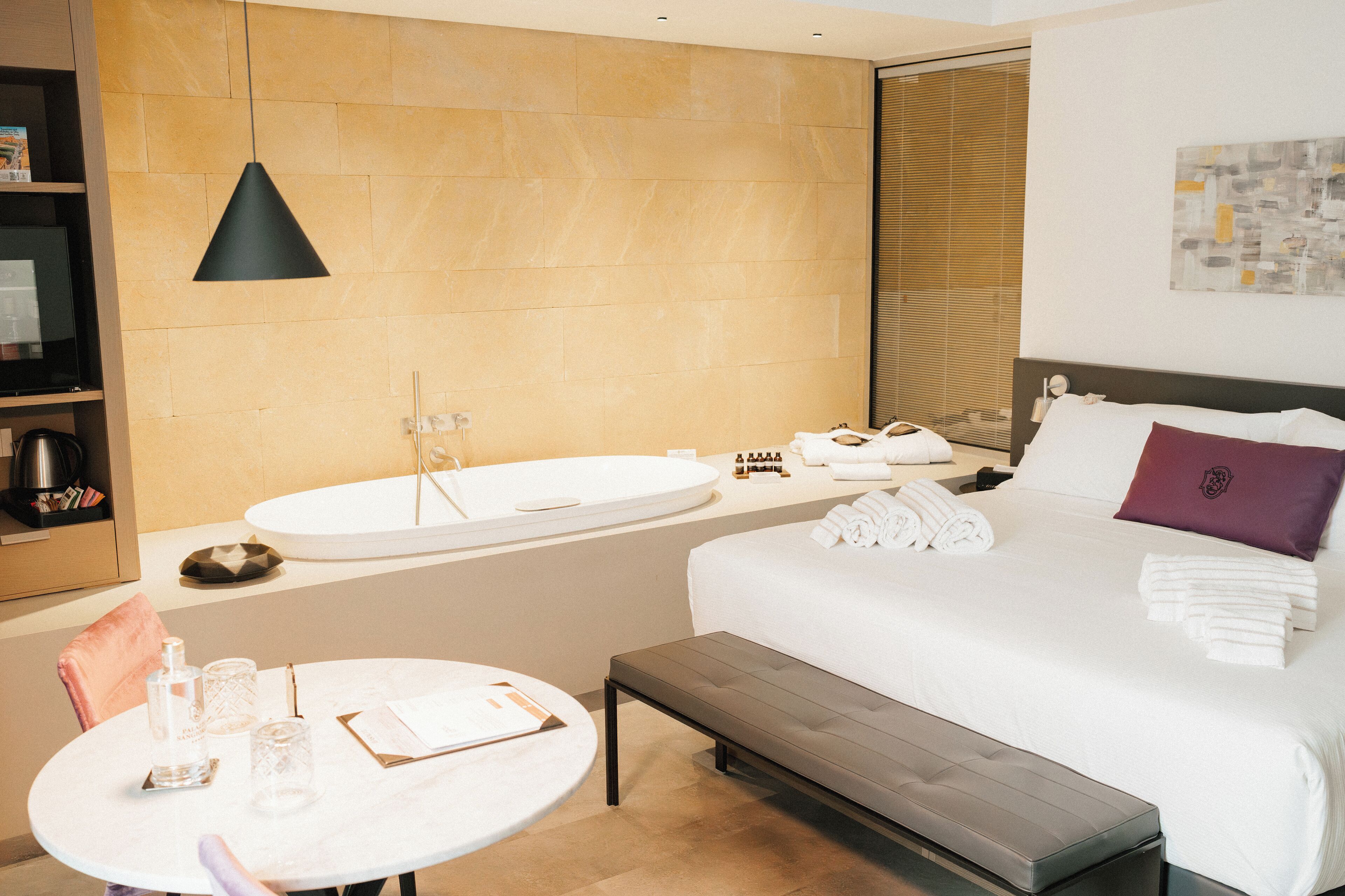A modern hotel room with a large bed, a round table with a chair, a small sink, and a bench.