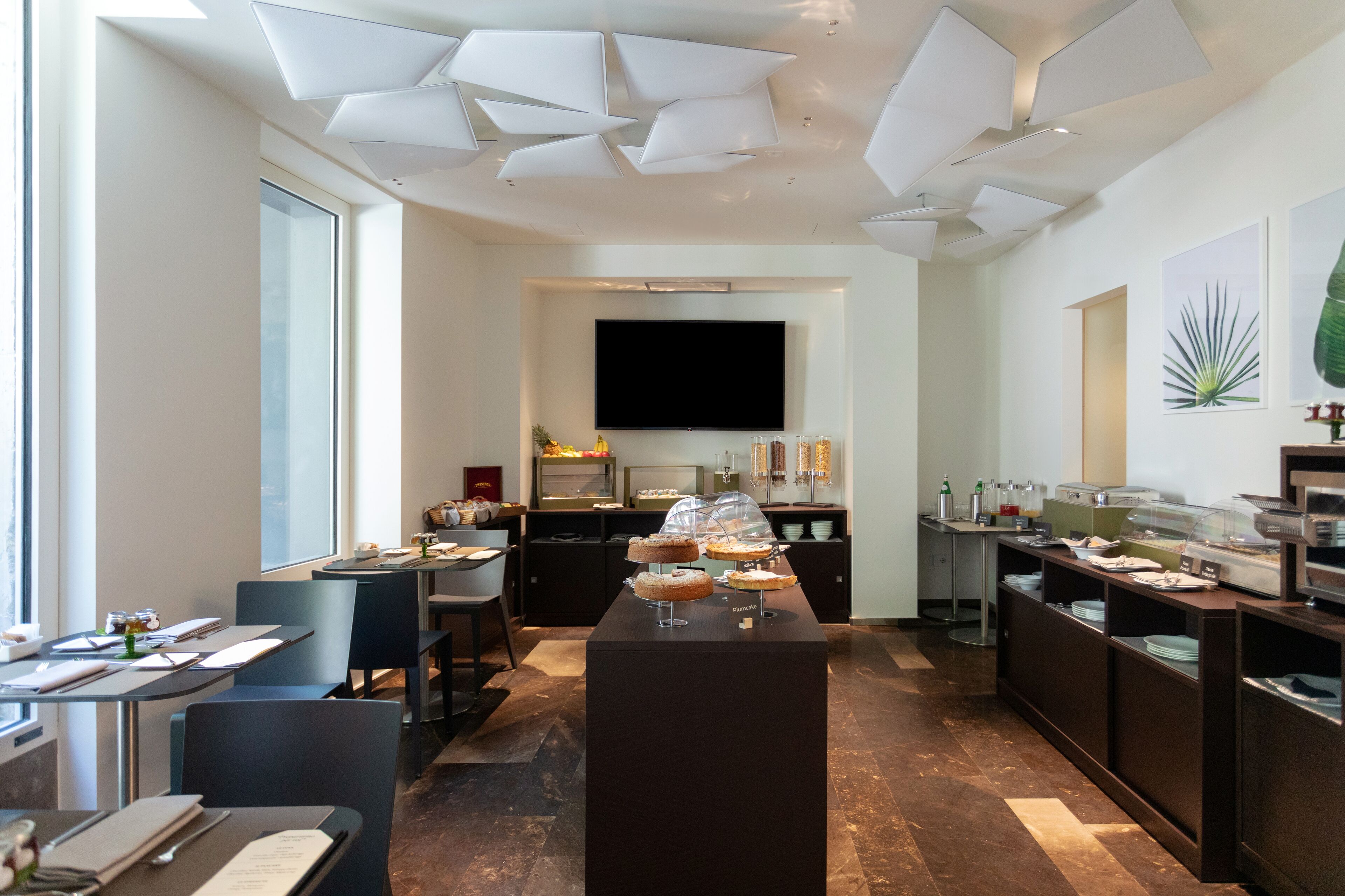 A modern hotel dining area with a central serving station, multiple tables, and a large flat-screen TV.