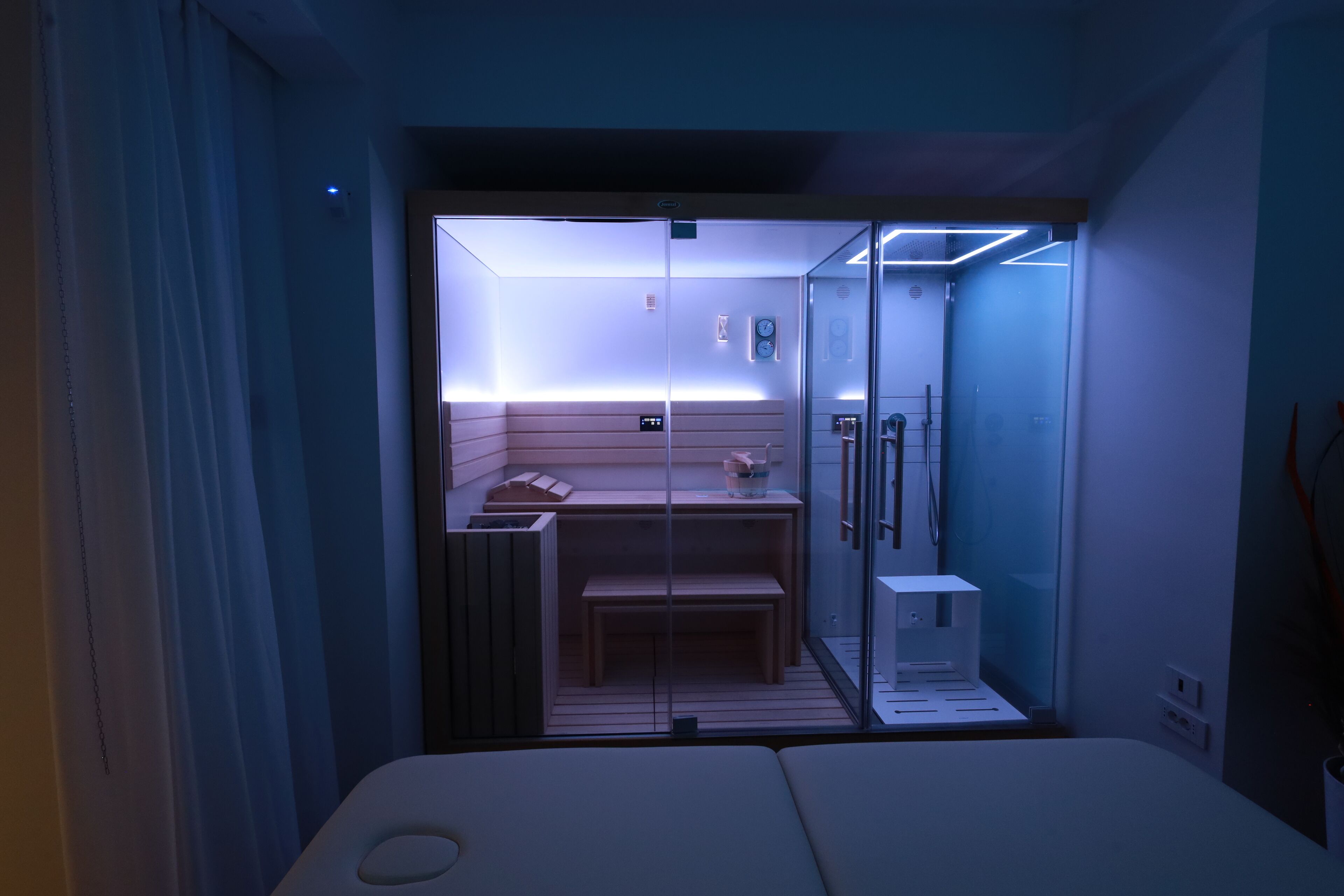 A compact, modern room with a bed, a small seating area, and a glass partition separating the sleeping area from a bathroom with a shower and a toilet.