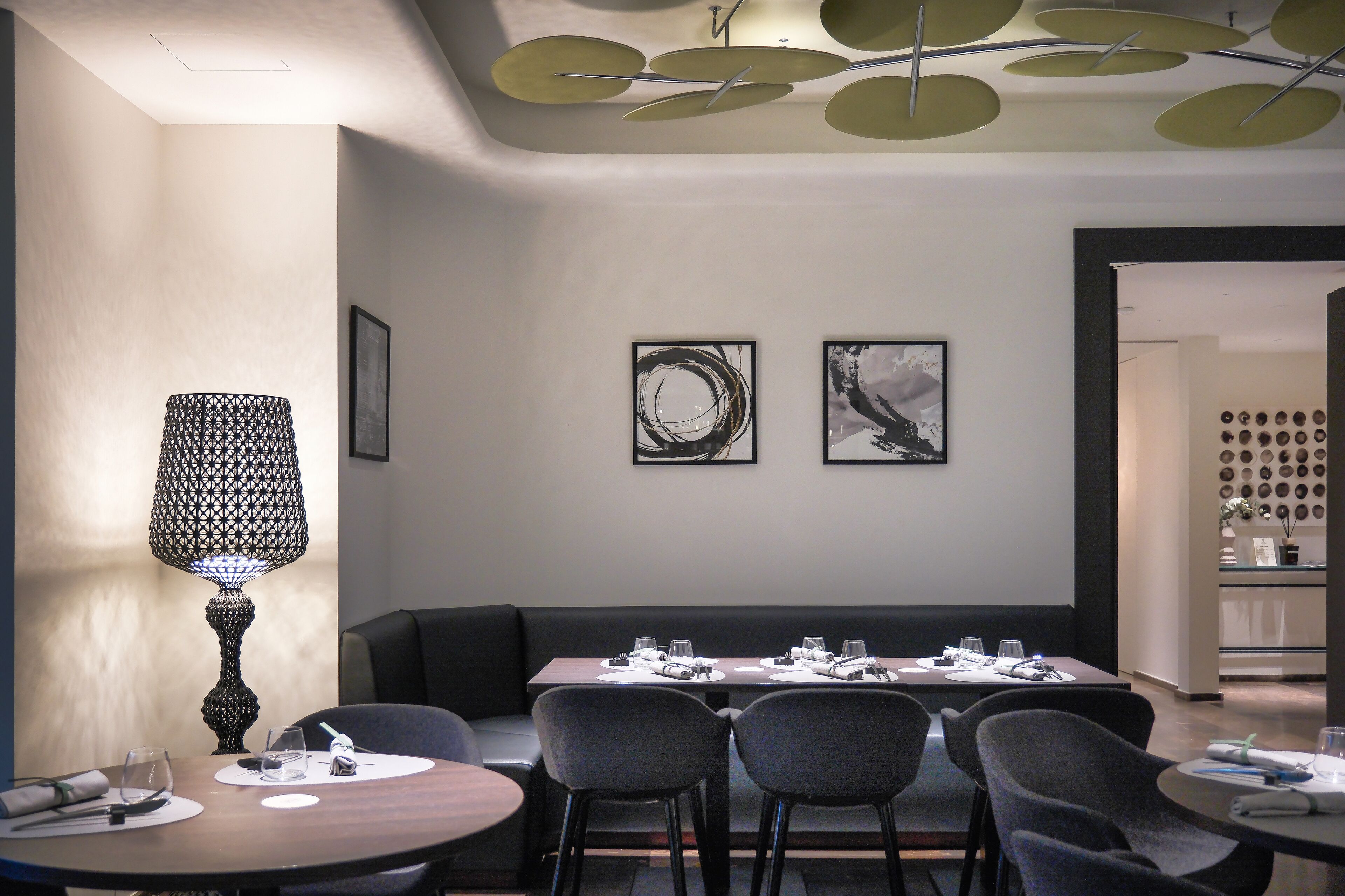 A modern dining area with a round table, a rectangular table, and a sofa. There are framed artworks on the wall and a decorative ceiling light.