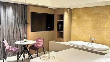 Una habitación de hotel moderna con una cama grande, una mesa redonda con sillas, un televisor, un minibar y una bañera.
