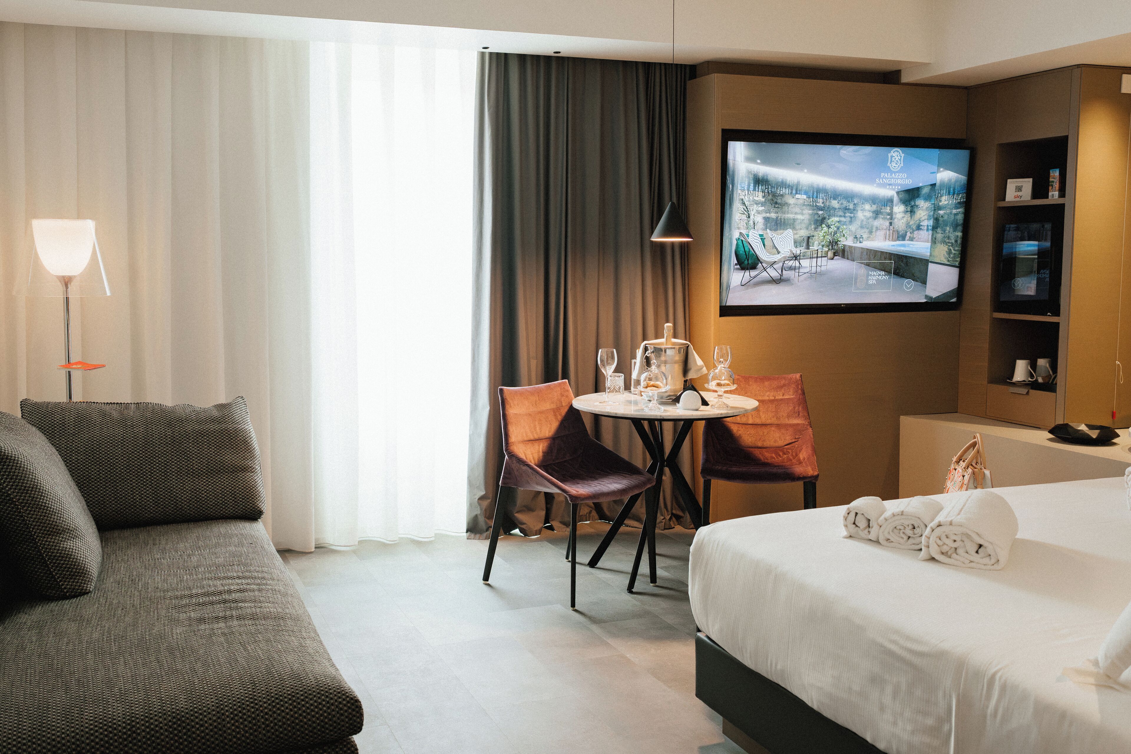 Une chambre d’hôtel comprenant un lit, un canapé, une table à manger avec des chaises, un téléviseur fixé au mur et une fenêtre avec des rideaux.