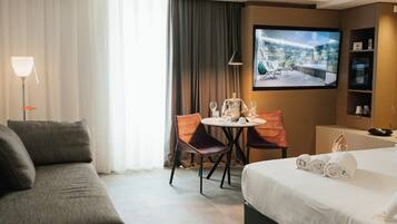 Une chambre d’hôtel comprenant un lit, un canapé, une table à manger avec des chaises, un téléviseur fixé au mur et une fenêtre avec des rideaux.