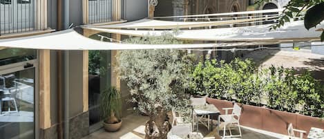 An outdoor café area with tables and chairs, surrounded by greenery and a stone fountain.
