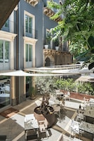 An outdoor café area with tables and chairs, surrounded by greenery and a stone fountain.