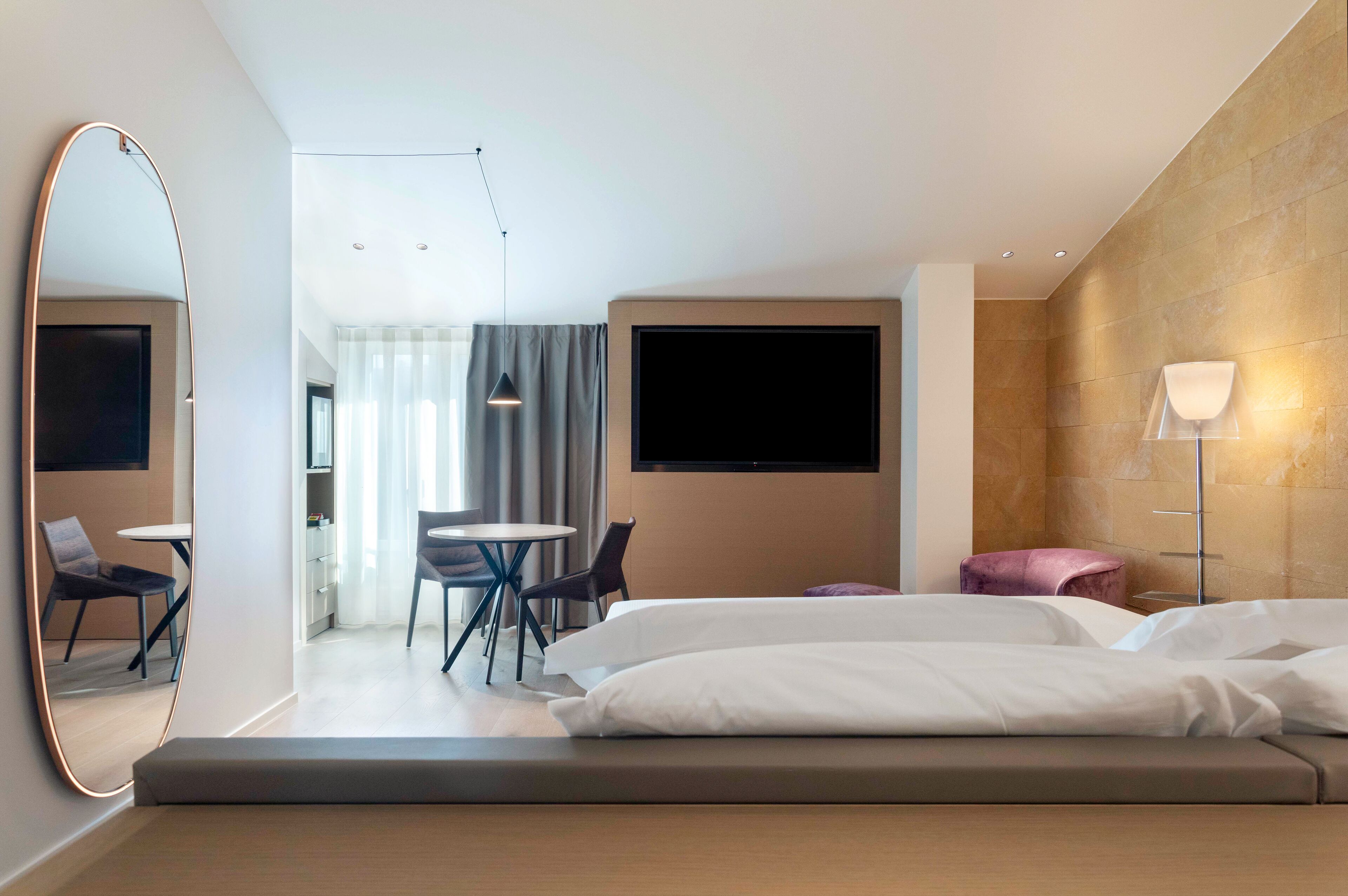 A modern hotel room with a large bed, a desk with chairs, a dining area, and a TV mounted on the wall.