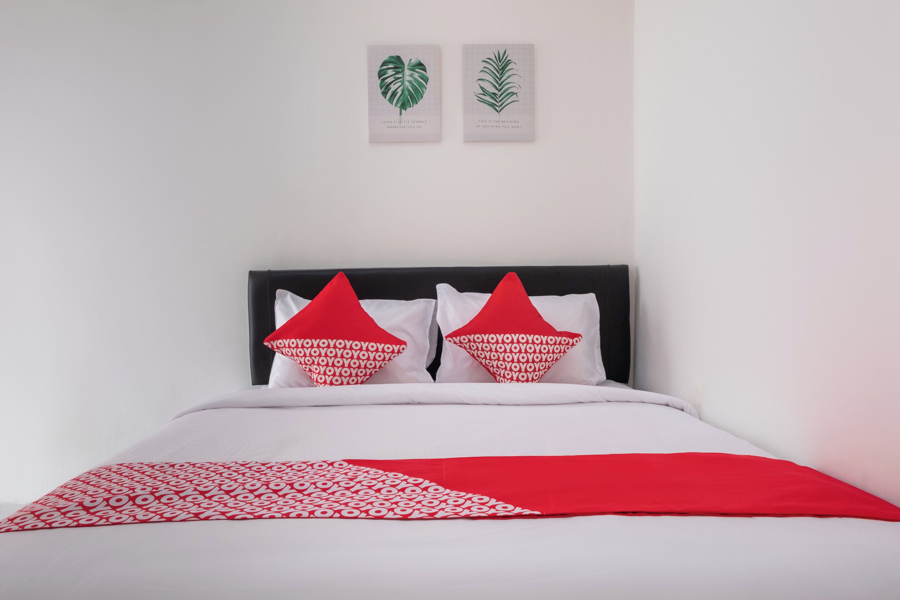 Ein Bett mit weißroter Bettdecke, zwei rote Kissen mit weißer Schrift sowie zwei gerahmte Pflanzenbilder an der Wand.