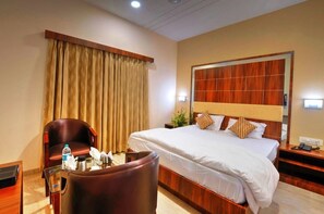 Habitación de hotel con una cama grande, cabecera de madera, sillón de cuero, mesita de noche con lámpara y una ventana con cortinas.
