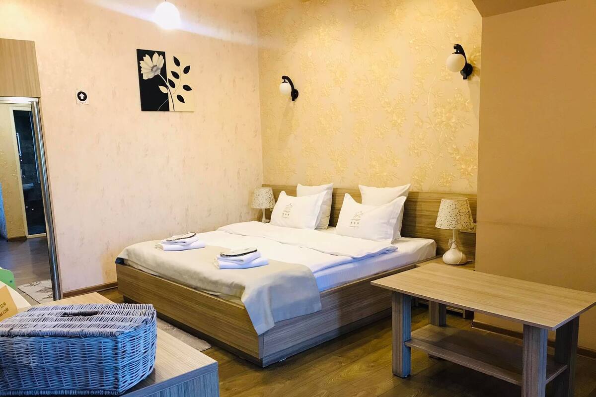 A hotel room with a wooden bed, white bedding, a small bedside table, and a wall-mounted lamp.