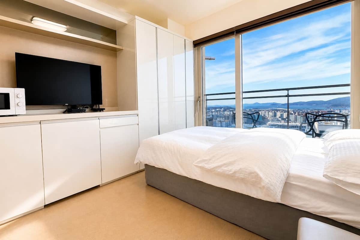 Un dormitorio moderno con cama, televisor y vistas a un paisaje urbano a través de amplios ventanales.