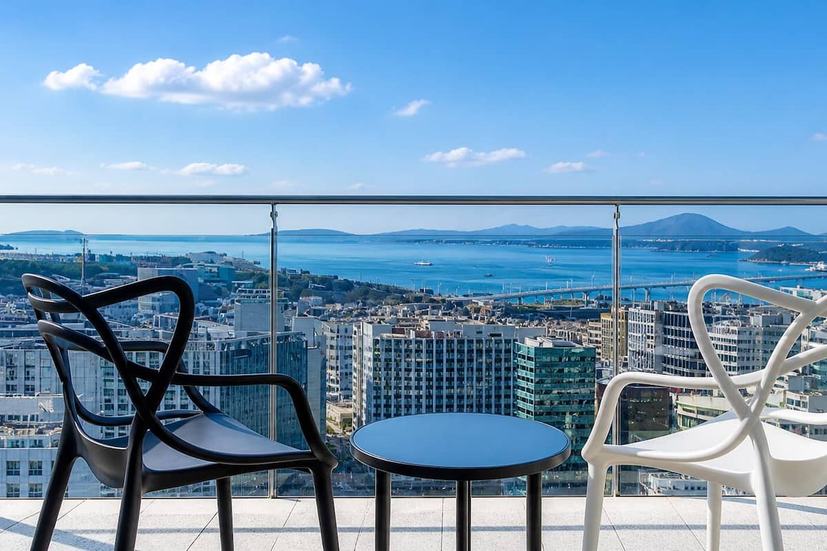 Un balcón con dos sillas y una mesa, con vistas a un paisaje urbano que incluye edificios y una masa de agua.