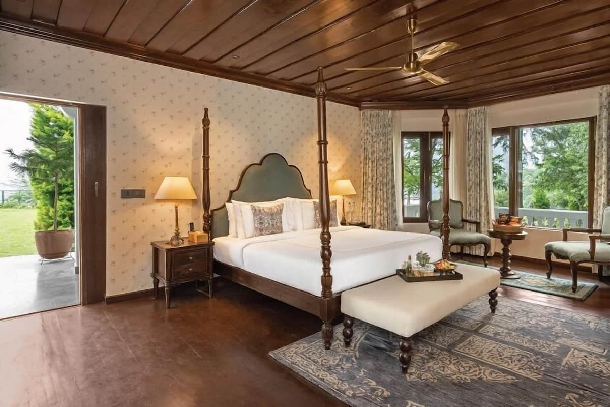 A four-poster bed with a wooden headboard, a bedside table with a lamp, a chair, and a view of greenery outside.